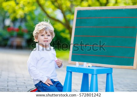 Adorable funny little kid boy at blackboard practicing counting and math, outdoor school or nursery. Child having fun with learning. Back to school concept.