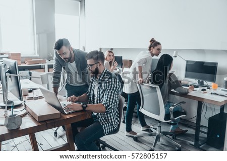 Achieving best results. Two confident young men looking at laptop monitor while their colleagues working in the background Royalty-Free Stock Photo #572805157