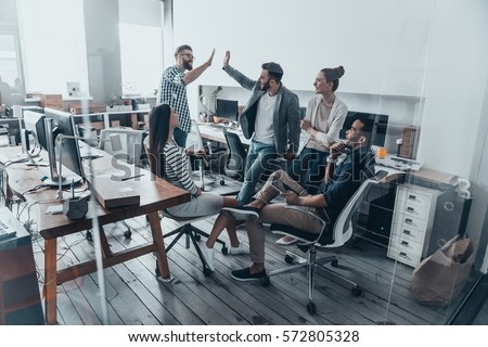 High-five for success! Two cheerful young business people giving high-five while their colleagues looking at them and smiling  Royalty-Free Stock Photo #572805328