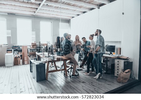 Team at work. Full length of young creative people in smart casual wear having a brainstorm meeting while standing behind the glass wall in the creative office Royalty-Free Stock Photo #572823034