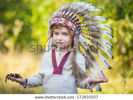 boy and spider on his shoulder, arachnid, blond boy in traditional American headdress, child and tarantula, the boy's face with freckles, headdress with eagle feathers, greenery background.