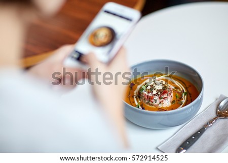 food, new nordic cuisine, technology, eating and people concept - woman with smartphone photographing bowl of vegetable pumpkin-ginger soup with goat cheese and tomato salad with yogurt at restaurant