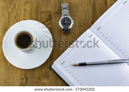 On a wooden surface lies an open diary with a pen, a watch with a bracelet and a cup of coffee costs