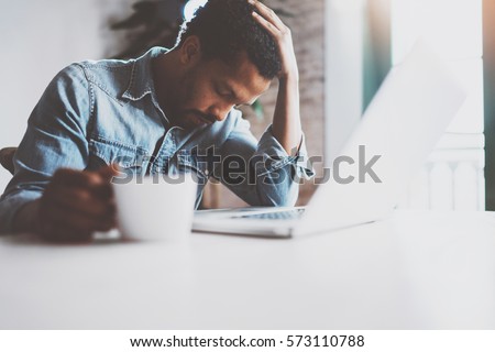 Tired young African man using laptop while sitting at the table on a sunny morning.Concept of people working hard home.Blurred background,flare effect Royalty-Free Stock Photo #573110788