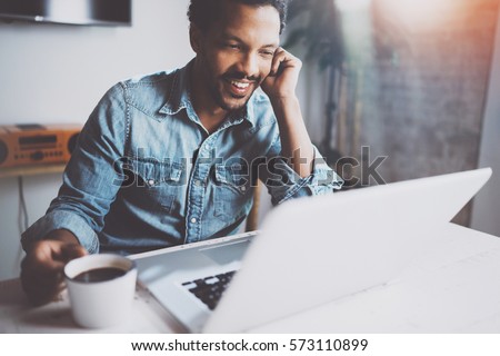 Happy bearded African man making video conversation via modern laptop with partners while holding white cup black coffee at home.Concept of young business people.Blurred background,flares effect Royalty-Free Stock Photo #573110899