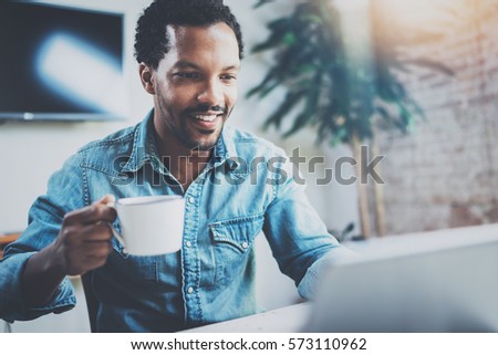 Smiling attractive African man making video conversation via digital tablet with business partners while sitting in sunny meeting room.Concept of happy coworking people.Blurred background,flares