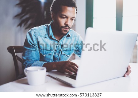 Pensive bearded African man working at home while sitting the wooden table.Using modern laptop for new job search.Concept of young people work mobile devices.Blurred background.Crop Royalty-Free Stock Photo #573113827