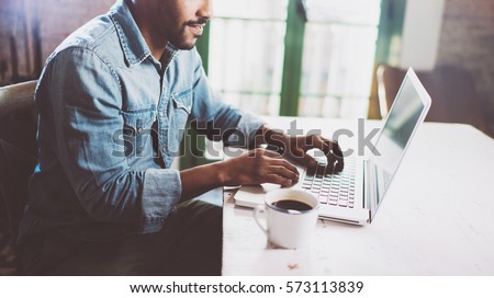 Smiling bearded African man using laptop at home while sitting the wooden table.Guy is typing on the notebook keyboard.Concept of young people work mobile devices.Blurred window background,crop Royalty-Free Stock Photo #573113839