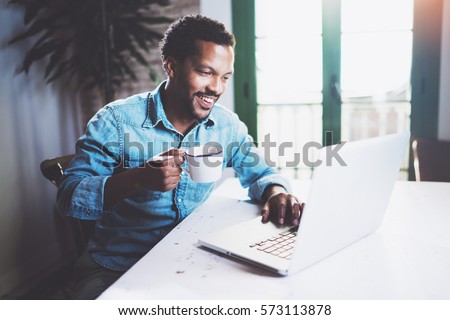 Smiling bearded African man working at home while sitting the wooden table.Using modern laptop for new job search.Concept of young people work mobile devices.Blurred background Royalty-Free Stock Photo #573113878