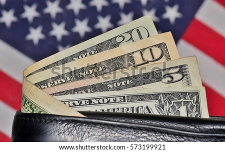 Partial leather wallet displaying US cash dollars with American flag blurred in background. USA economics and finance concept.
