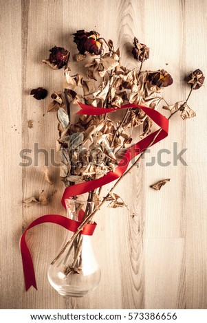 Dried bouquet of roses in glass vase wrapped with red ribbon, fallen on a wooden floor.