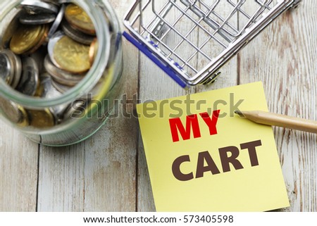 Words My Cart written on sticky note with pencil, mini shopping cart and glass jar full with coins on wooden background. Conceptual.