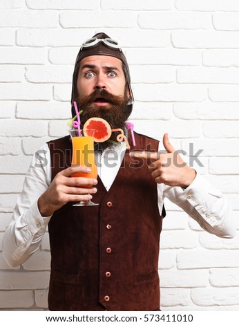 handsome bearded aviator man with long beard and mustache on surprised face holding glass of alcoholic cocktail in vintage suede leather waistcoat with hat and glasses on white brick wall background