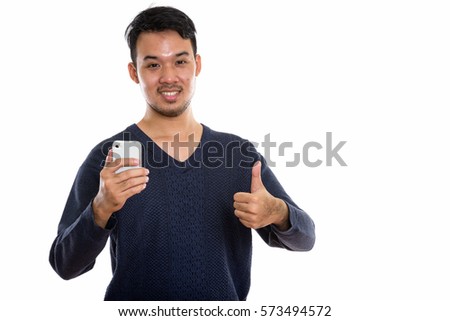 Studio shot of young happy Asian man smiling while holding mobile phone and giving thumb up