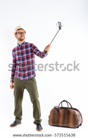 Full body shot of happy young man smiling while holding selfie stick and taking selfie picture with mobile phone ready for vacation