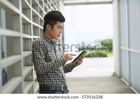 Profile view of handsome young man in stylish clothes leaned back to white wall and surfing net on digital tablet, waist-up portrait