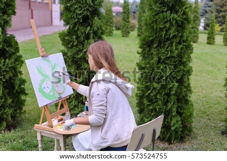 Pretty attractive artist begins to painting which depicts  colorful butterfly, sitting with back to photographer and behind wooden easel, holds palette smeared in paint in large park green day outdoor