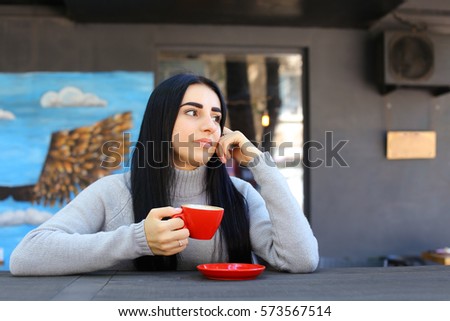 Charming young female girl smiling, holding red mug in hand, looks at camera, looks away, drinking coffee at wooden table on gray background panoramic windows and walls decorated with pictures. Girl