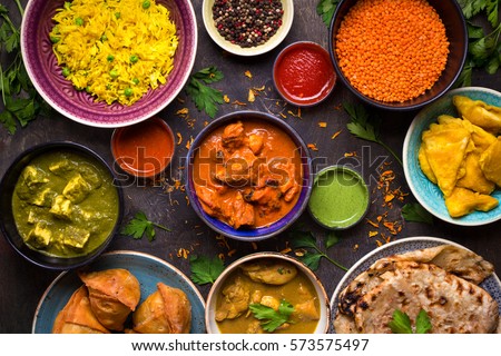 Assorted indian food on dark wooden background. Dishes and appetizers of indian cuisine. Curry, butter chicken, rice, lentils, paneer, samosa, naan, chutney, spices. Bowls and plates with indian food Royalty-Free Stock Photo #573575497