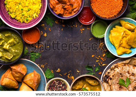 Assorted indian food on dark wooden background. Dishes of indian cuisine. Curry, butter chicken, rice, lentils, paneer, samosa, naan, chutney, spices. Space for text. Bowls and plates with indian food Royalty-Free Stock Photo #573575512
