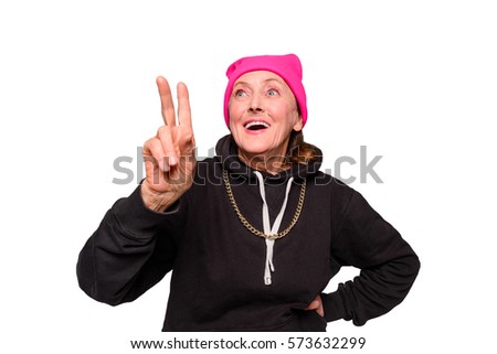 Granny shows a peace sign. Crazy grandmother in a black hoodie with a long gold chain and a vivid pink hat. Studio shot of an old woman in a lifted mood over white background.
