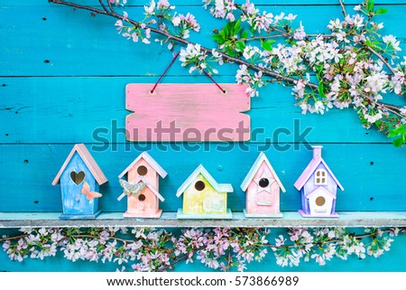 Blank pink sign hanging over colorful birdhouses with butterfly on shelf by spring tree flowers on antique rustic teal blue wooden background; springtime background with painted wood copy space