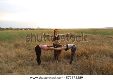 Girls perform postures or asanas of yoga, posing for photographer to take advertising pictures for sports women's clothing catalog, meditate outdoors in wide field. Girl with shoulder-length hair