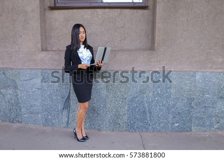 Promising young entrepreneur beautiful woman uses laptop for work, make appointment, solves important business issues and and stands right in all growth against the background of gray wall of business