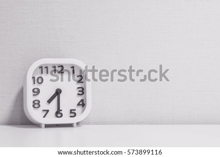 Closeup white clock for decorate show half past seven or 7:30 a.m. on white wood desk and cream wallpaper textured background in black and white tone with copy space