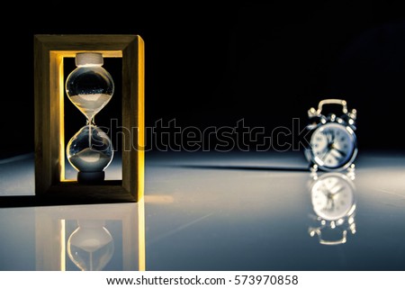 low key photography of an hourglass suggesting the passage of time with a little clock on background in darkness. (colored version)