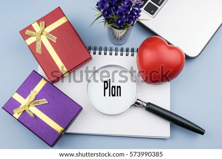 PLAN wording in magnifier glass with small gift, red heart, notebook and laptop on desk. Business, finance, economy concept