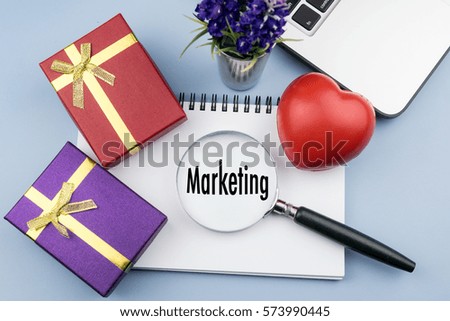 MARKETING wording in magnifier glass with small gift, red heart, notebook and laptop on desk. Business, finance, economy concept
