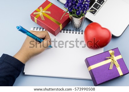 Close up of hand writing on empty notebook, with small gift, red heart and laptop on desk. Wishes, education, business, concept
