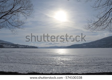 Sunshine through clouds reflecting on icy lake with tree branches in foreground. Cloudy sky, horizon and mountains in background.