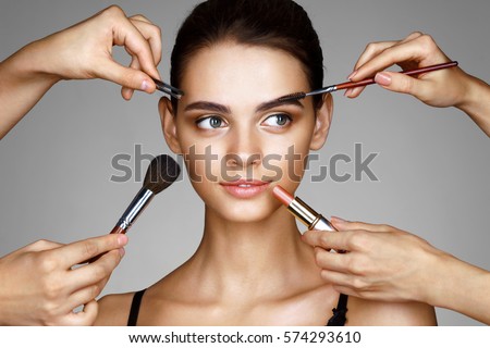 Beautiful girl surrounded by hands of makeup artists with brushes and lipstick near her face. Photo of happy woman on gray background. Grooming himself
