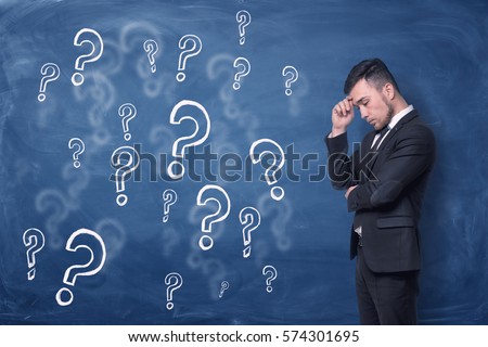 A businessman is standing thinking near blue blackboard background which is full of question marks. Puzzlement. Man thinking.