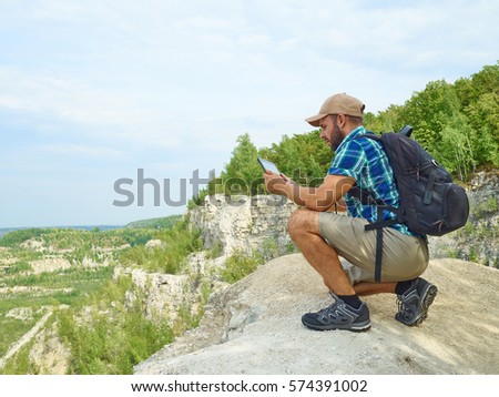 Man Tourist Uses Tablet Computer Sitting on Edge of a Cliff In Mountains.
