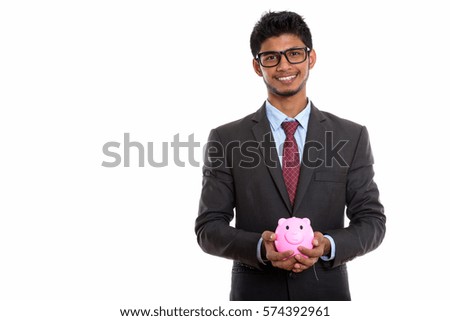 Studio shot of young happy Indian businessman smiling while holding piggy bank