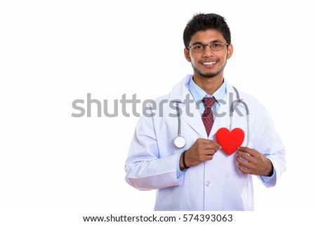 Studio shot of young happy Indian man doctor smiling while holding red heart against chest