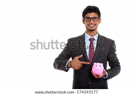 Studio shot of young happy Indian businessman smiling while holding and pointing at piggy bank