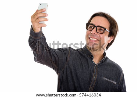 Studio shot of young happy Persian man smiling while taking selfie picture with mobile phone