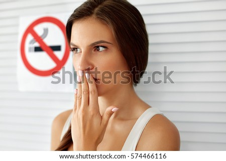 No Smoking. Portrait Of Beautiful Young Woman Closing Her Mouth With Hand Standing Near No Smoking Sign On Wall In Non-Smoking Area, Smoke Free Zone. High Resolution 