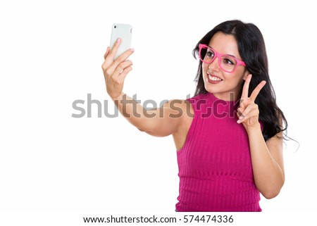 Studio shot of young happy Spanish woman smiling while taking selfie picture with mobile phone and giving peace sign