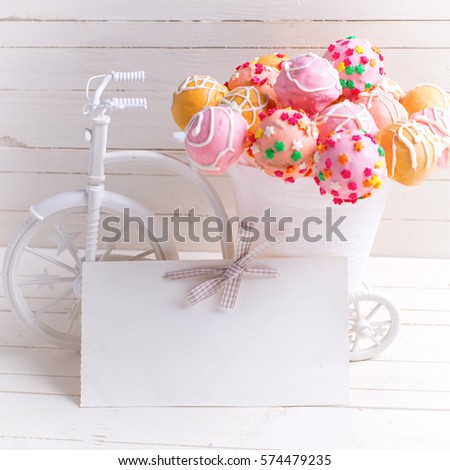 Empty tag and bright cake pops  in decorative bicycle on white wooden background. Selective focus. Place for text. Square image.
