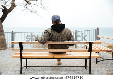 Back view picture of young african man wearing hat and scarf sitting on a bench on the street.