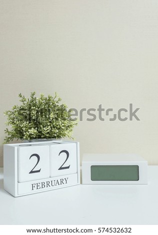 White wooden calendar with black 22 february word with clock and plant on white wood desk and cream wallpaper textured background in selective focus at the calendar
