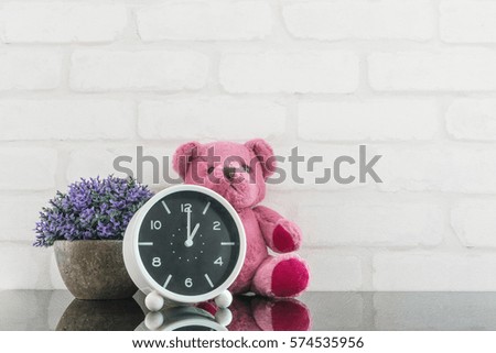 Closeup black and white alarm clock for decorate in 1 o'clock with bear doll and plant on black glass table and white brick wall textured background with copy space