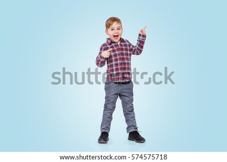 Full length portrait of a little boy standing and pointing finger away isolated over blue background