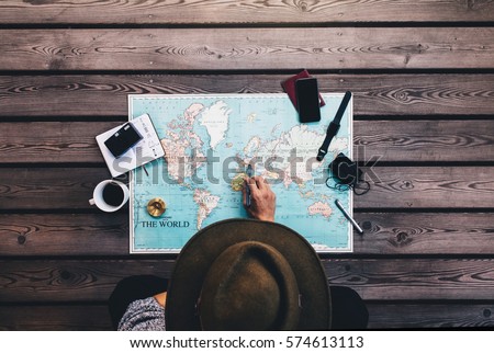 Tourist pointing at Europe on world map surrounded with binoculars, compass and other travel accessories. Man wearing brown hat planning his tour looking at the world map. Royalty-Free Stock Photo #574613113