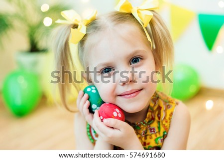 blond, blue-eyed pretty girl smiling ponytail hair in yellow dress 4-5 years with Easter eggs in a room on the background of yellow design studio